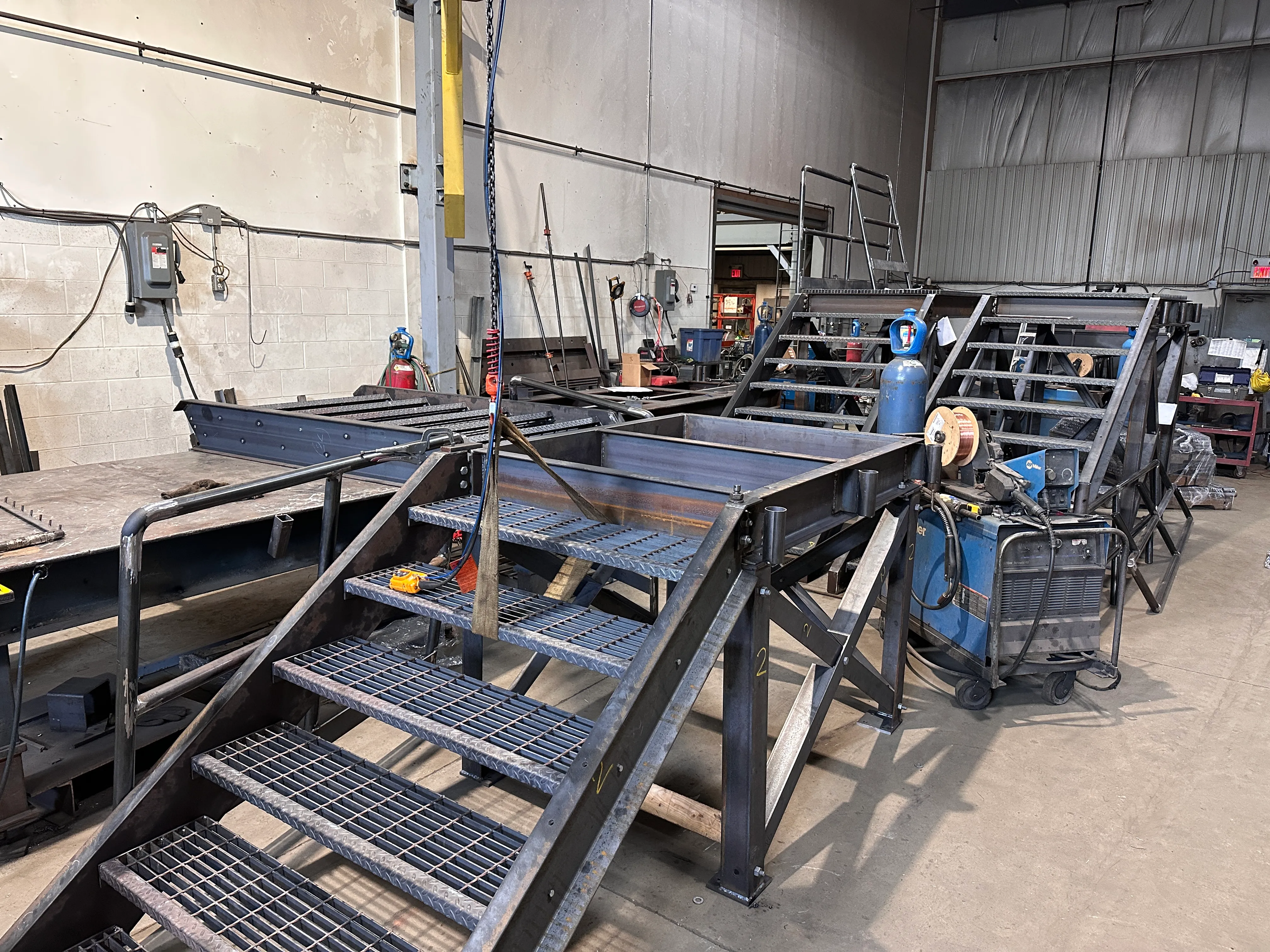 Fabricated steel stairs and platforms mid-build in the shop beside welding equipment.